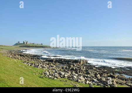 Cielo azzurro sopra il castello di Dunstanburgh in Northumberland con onde che si infrangono sulla costa rocciosa. Foto Stock