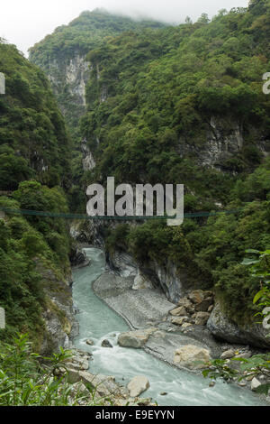 Scenario paesaggistico di lussureggianti e ripide montagne, gola profonda e una sospensione ponte su un fiume roccioso presso il Parco Nazionale di Taroko Foto Stock