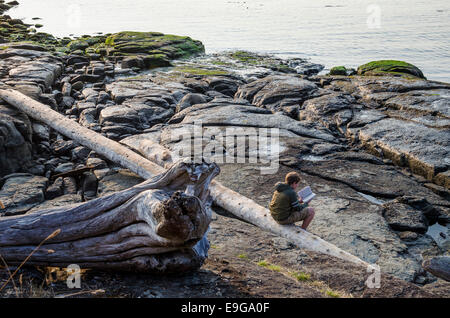 Ragazzo lettura libro, seduto su driftwood log, Ruckle Parco Provinciale, sale spring Island, British Columbia, Canada Foto Stock