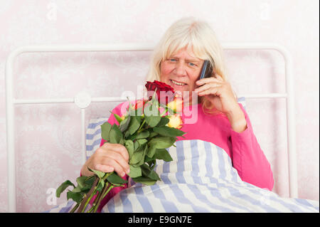 Donna felice in rosa pigiama seduta nel letto con rose e parlando al telefono Foto Stock