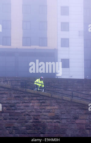 Lavoratore in abiti ad alta visibilità che cammina accanto a un muro di pietra nella nebbiosa Nottingham, con edifici moderni che svaniscono nella nebbia Foto Stock