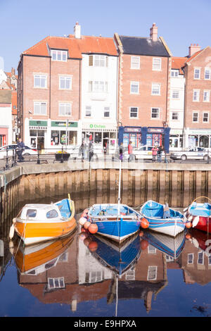 Piccole e colorate barche ormeggiate nel porto di Whitby con riflessi luminosi e storici edifici sul lungomare con la calda luce del mattino Foto Stock