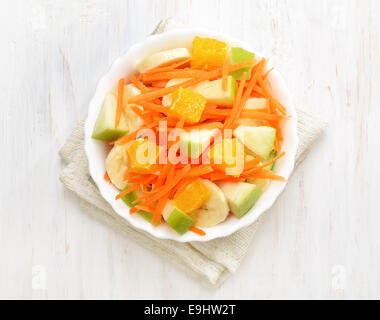 Frutta con Insalata di carote, mele, banane e fettine di arancia in una terrina bianco sul tavolo di legno, vista dall'alto Foto Stock