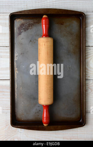 Angolo alto ingrandimento di un vecchio legno perno di rotolamento con maniglie rosse su un ben utilizzati cookie foglio. Il pan è su un tavolo rustico Foto Stock