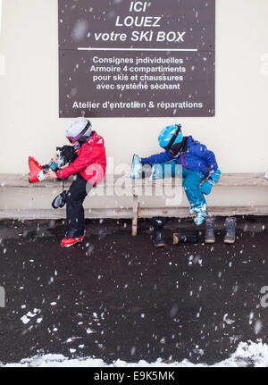 Piccolo Ragazzo e ragazza di abbigliamento sci caschi e occhiali seduta su una panchina mettendo su scarponi da sci con caduta di neve Foto Stock