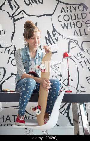 Lunghezza intera della ragazza con lo skateboard seduto sul tavolo da studio a casa Foto Stock