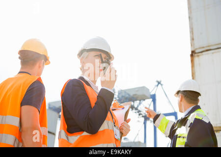 Supervisore femmina utilizzando un walkie-talkie mentre i lavoratori di discutere in background in cantiere Foto Stock