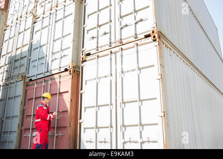 Lavoratore di sesso maschile di ispezionare contenitori di carico durante la scrittura sulla clipboard in cantiere di spedizione Foto Stock