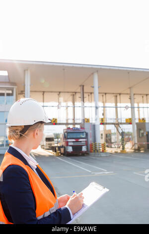 Lavoratore di sesso femminile che la scrittura sulla clipboard mentre guardando i camion di entrare nel cantiere di spedizione Foto Stock