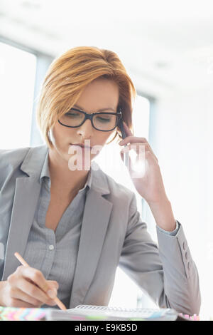 Giovane imprenditrice su chiamata mentre è in corso la scrittura alla scrivania in ufficio Foto Stock