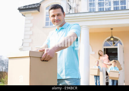 Ritratto di fiducioso uomo che porta una scatola di cartone mentre casa in movimento con la famiglia in background Foto Stock