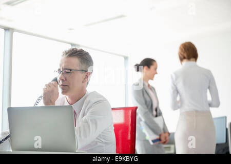 Imprenditore parlando al telefono con i colleghi in background in ufficio Foto Stock