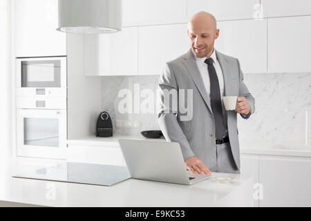 Adulto medio imprenditore avente caffè mentre utilizzando laptop in cucina Foto Stock