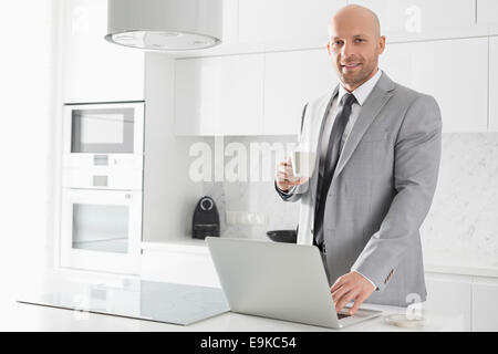 Ritratto di fiducioso metà imprenditore adulti aventi caffè mentre utilizzando laptop in cucina Foto Stock