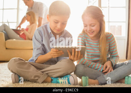 Felice fratelli utilizzando digitale compressa sul pavimento con i genitori in background Foto Stock