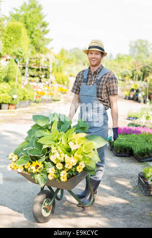 Ritratto a figura intera di felice giardiniere carriola spinta con piante in giardino Foto Stock