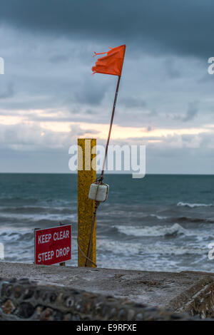 Tenersi a distanza ripida discesa segno di avvertimento sulla spiaggia accanto a una bandiera arancione contro un sfondo tempestoso Foto Stock