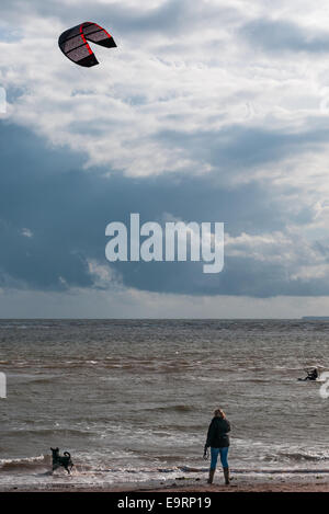 Un kite surfer in una giornata ventosa a Exmouth nel Devon, Regno Unito. Buone condizioni per il kite surf. Una donna e il suo cane guarda dalla spiaggia Foto Stock