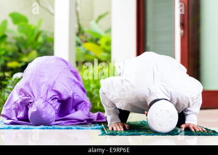 Musulmani asiatici giovane, uomo e donna, pregando in casa seduti sul tappeto di preghiera nella loro casa di fronte al giardino tropicale Foto Stock