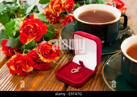 Gioielli in oro in velluto rosso scatola, tazze di tè e bouquet di rose sul tavolo di legno Foto Stock