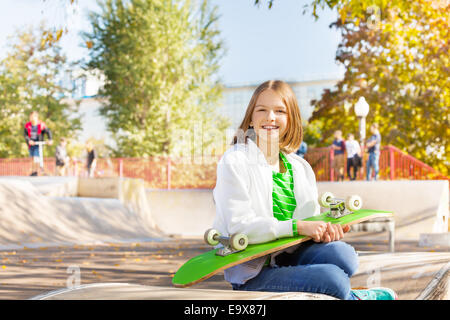 Sorridente ragazza trattiene sullo skateboard, siede nel parco giochi Foto Stock