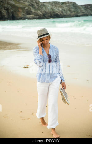 Donna matura in chat sullo smartphone mentre per passeggiare sulla spiaggia, Camaret-sur-mer, Bretagna Francia Foto Stock