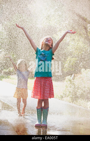 Due ragazze con i bracci aperti in piedi in uno spruzzo di acqua su strada Foto Stock
