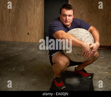 Ritratto di giovane maschio weightlifter squatting con atlas palla in palestra Foto Stock