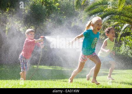 Tre bambini in giardino che si rincorrono con soffione di erogazione dell'acqua Foto Stock