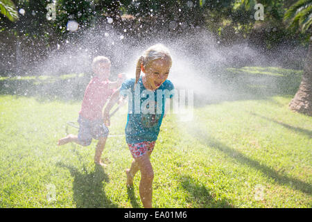 Ragazzo in esecuzione dopo la ragazza in giardino con sprinkler acqua Foto Stock