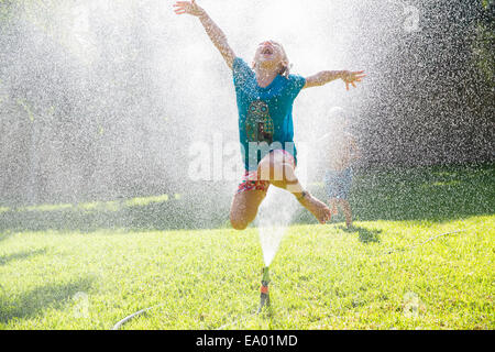 Ragazza saltando soffione di erogazione dell'acqua in giardino Foto Stock