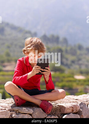 Ragazzo seduto sul muro di pietra guardando digitale compressa, Maiorca, SPAGNA Foto Stock