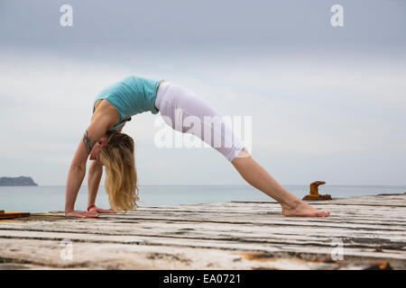 Metà donna adulta di piegarsi indietro la pratica dello yoga sul mare in legno pier Foto Stock