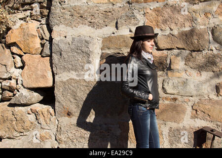 Elegante giovane donna in un nero giacca di pelle, alla moda cappello e sciarpa in piedi lateralmente contro un vecchio muro di pietra sotto il sole Foto Stock