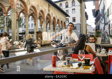 Giovane uomo Italiano in bicicletta si arresta per chattare con gli amici al cafè sul marciapiede opposto riposizionato loggia rinascimentale e il mercato delle pulci Foto Stock