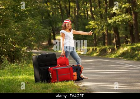 Giovani sollevatore-escursionista ragazza in piedi sul lato strada pomeriggio nella foresta con sacchetti Foto Stock