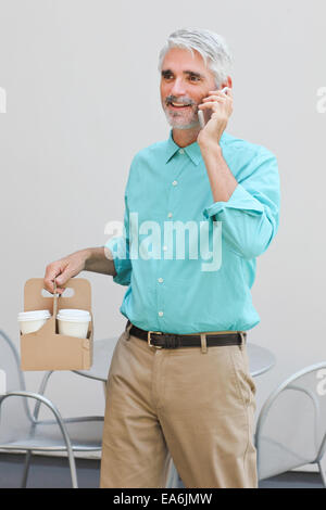 Imprenditore tenendo il contenitore con tazzine di caffè e parlando al telefono cellulare Foto Stock