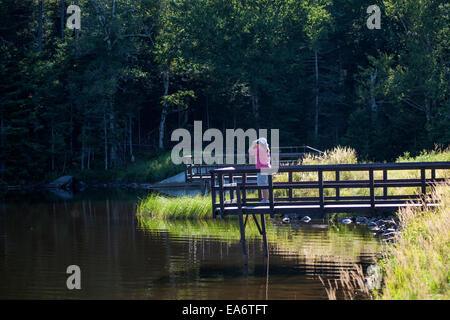 Una donna si erge su un dock e utilizza il binocolo per scorgere uccelli selvatici e altri animali selvatici in una zona umida nel New Hampshire, Stati Uniti d'America. Foto Stock