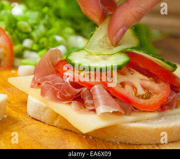 Donna mettendo mano cetriolo su bianco bloomer sandwich di pane di prosciutto, formaggio gruviera e fette di pomodoro Foto Stock