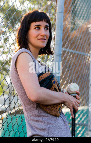 Donna che mantiene il baseball, il guanto e la mazza da baseball appoggiata contro la recinzione ad anelli Foto Stock