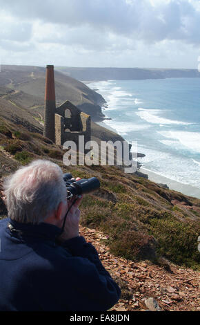 Uomo anziano che guarda al mare attraverso il binocolo su una scogliera sopra Wheal Coates dismesse Miniera di stagno Cappella Porth vicino a St Agnes su Foto Stock