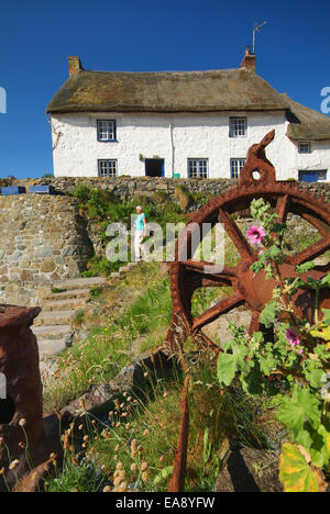 Arrugginimento ruota di metallo con dipinti di bianco cottage con tetto in paglia in background & lady passeggiando per le fasi di Cadgwith porto sul Lizar Foto Stock