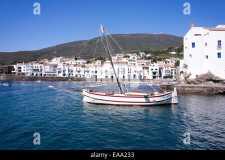 Spagna, tradizionale barca a vela in legno che galleggia sulle acque blu calme con una panoramica cittadina costiera e una collina sullo sfondo Foto Stock