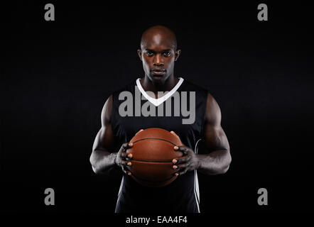 Ritratto di muscolare maschio giovane giocatore di basket in uniforme con una palla da basket che posano su sfondo nero Foto Stock