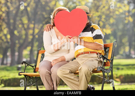 Coppia matura kissing dietro un cuore rosso seduto su una panchina nel parco Foto Stock