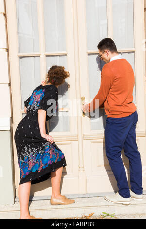 Uomo con occhiali in il maglione arancione con la donna cercando di aprire la porta mentre guardando attraverso la finestra e la tenda , giovane Foto Stock