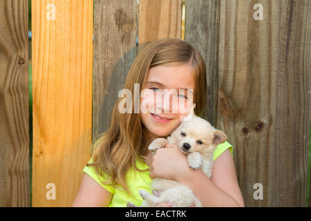 Blonde kid ragazza abbraccio di un cucciolo di cane felice chihuahua in backyard recinzione di legno Foto Stock