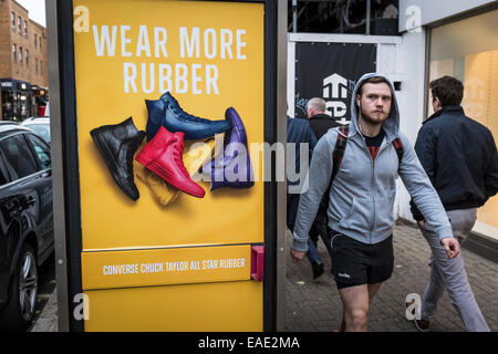 Uomo che indossa una felpa con cappuccio e pantaloncini camminare davanti a un annuncio colorati per scarpe di gomma. Foto Stock