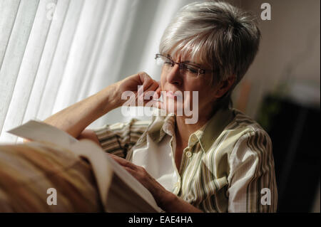 Senior donna seduta alla finestra e la lettura di un libro, Nord Reno-Westfalia, Germania Foto Stock