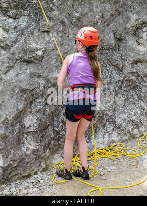 Sorridente ragazza giovane con occhiali da sole e arrampicata attrezzature compreso elmetto e il sistema di cavi in attesa di salire una parete di roccia. Foto Stock